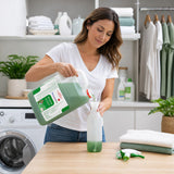 A woman in a white t-shirt and jeans pours green laundry detergent from a large plastic jug into a white spray bottle on a wooden table in a bright laundry room.