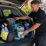 A man in a black polo shirt with a 'ProClean' logo stands by an open car trunk, organizing cleaning supplies including a large green bottle, blue gloves, a gray microfiber mop, and buckets.