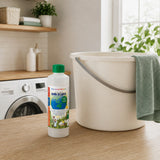 A bottle of laundry detergent from Down to Earst stands on a wooden countertop next to a large off-white plastic bucket with a gray handle and a green towel draped over the side. The background shows a bright, modern laundry room with a washing machine and shelves.